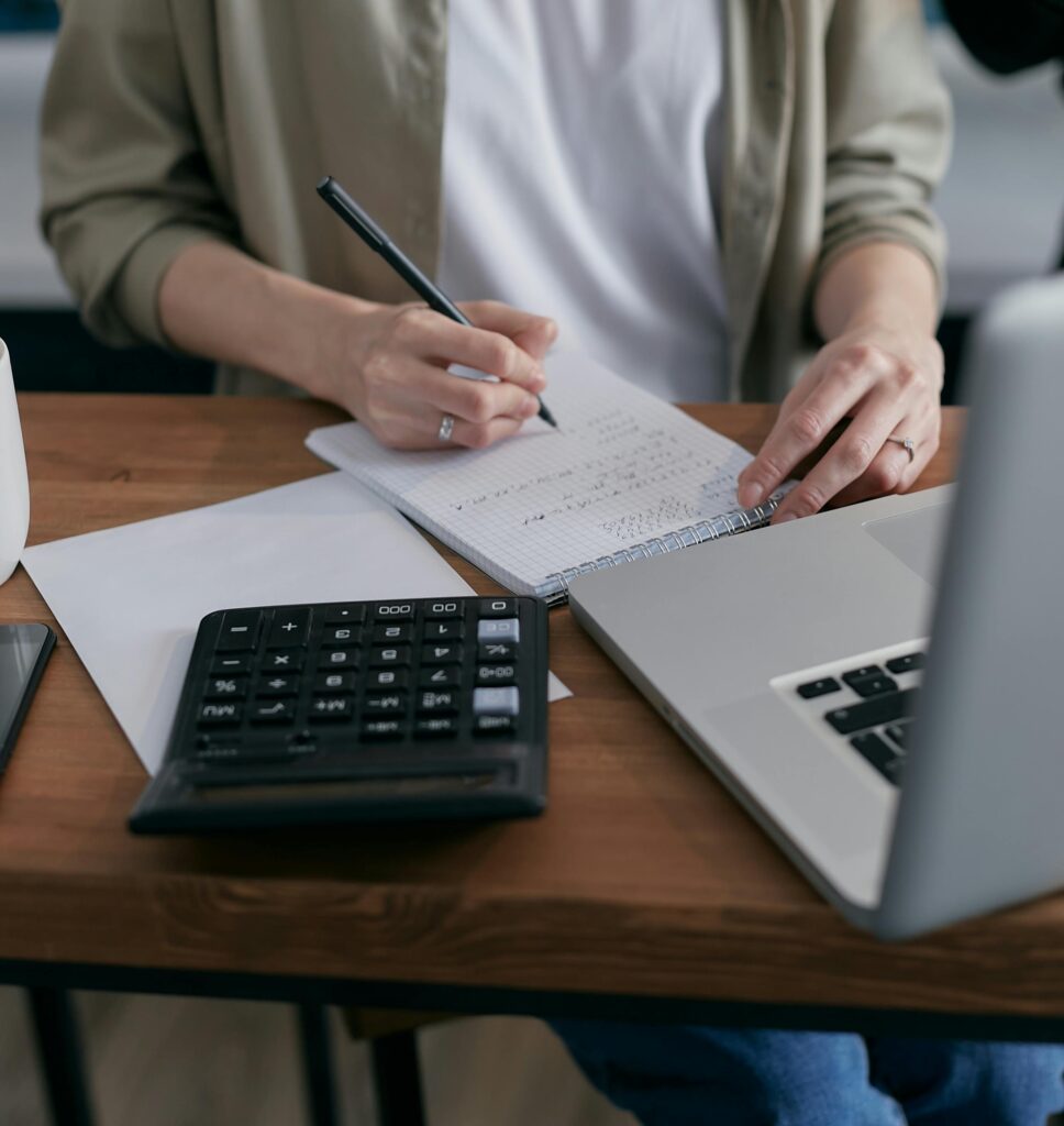 A woman writes financial calculations in a notebook, using a calculator and laptop at a wooden desk.